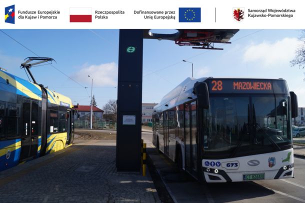 Bialy autobus miejski stojący pod ładowarką pantografową, obok niebiesko-żółty tramwaj